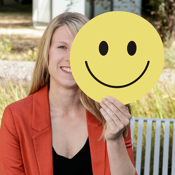 Die Beschäftigte Frau Deisler hält mit einem großen Smiley aus Papier die Hälfte ihres Gesichtes zu