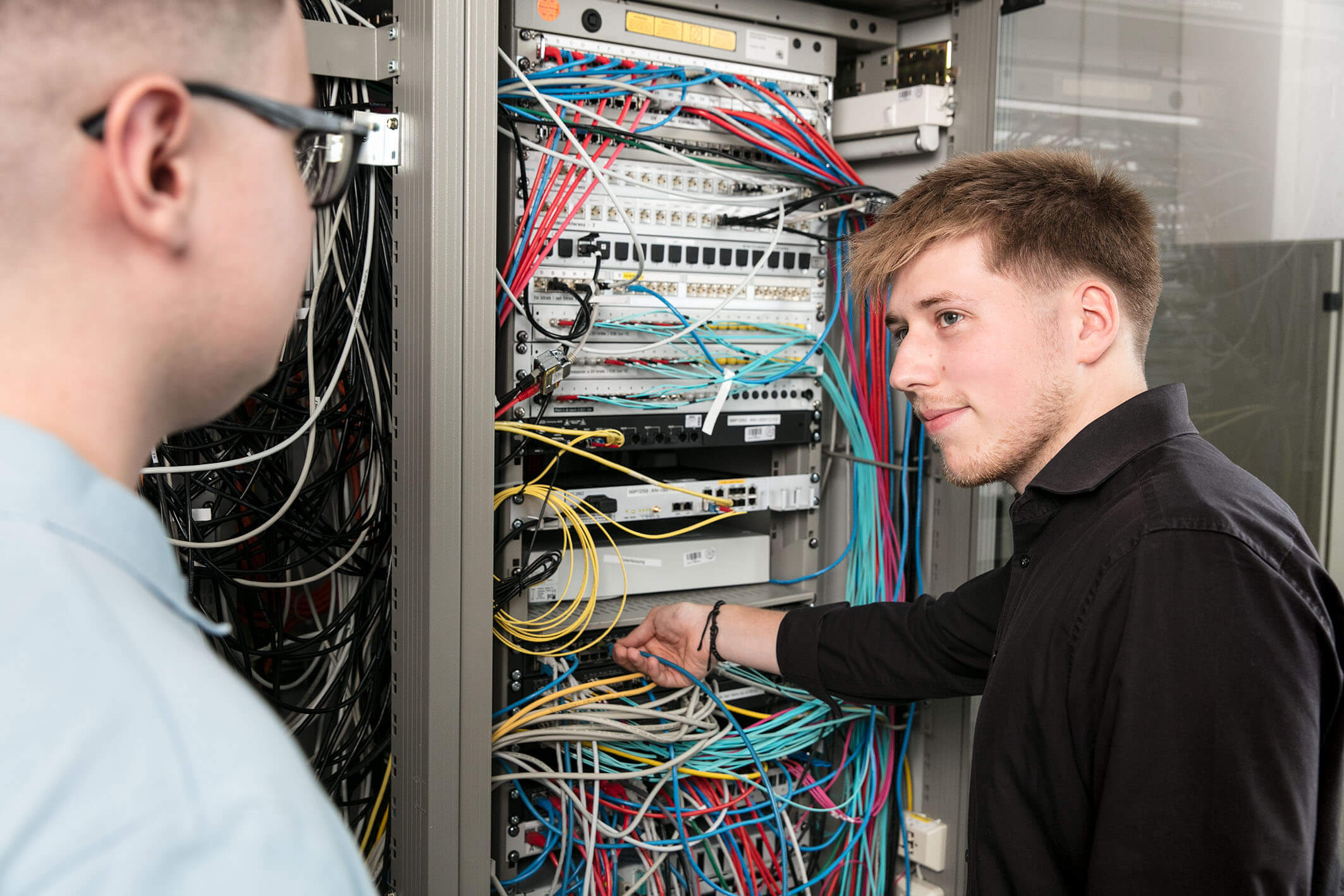 Zwei Auszubildende im Bereich Fachinformatik stehen vor einem geöffneten Serverschrank. Ein Auszubildender steckt ein Kabel um.